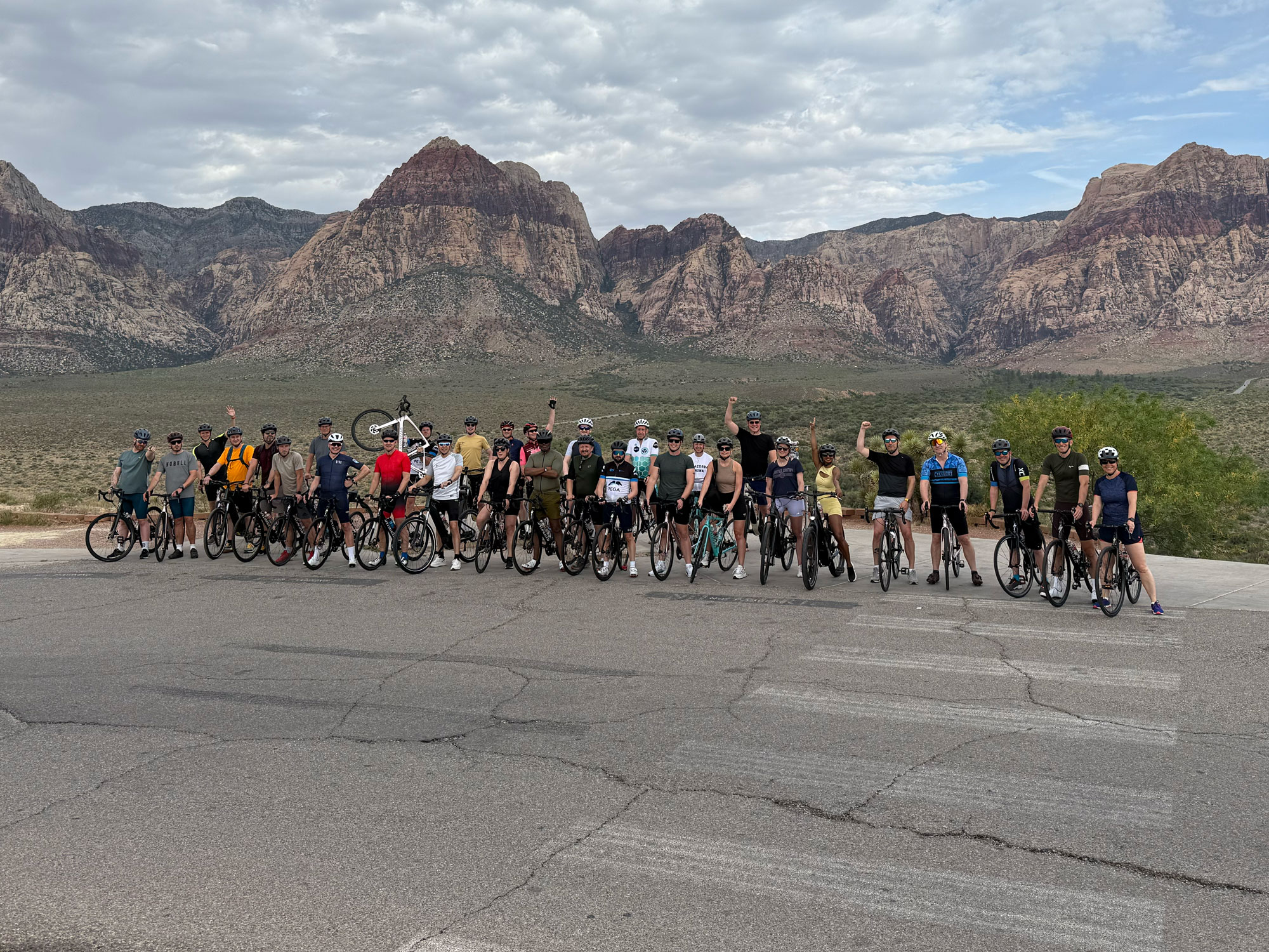 A group of cyclists standing in a line in front of a mountain range, triumphant after a gruelling team ride.